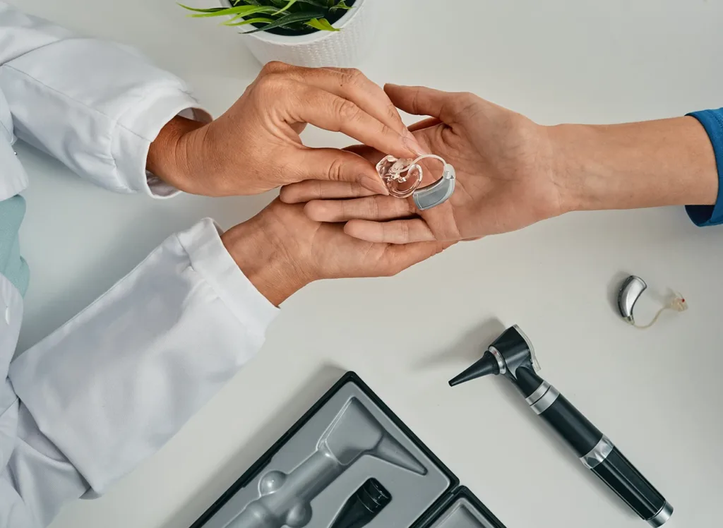 Top view of hearing care specialist putting a modern BTE hearing aid in patient hand at a hearing clinic; Hearing Aid clean and check appointments concept